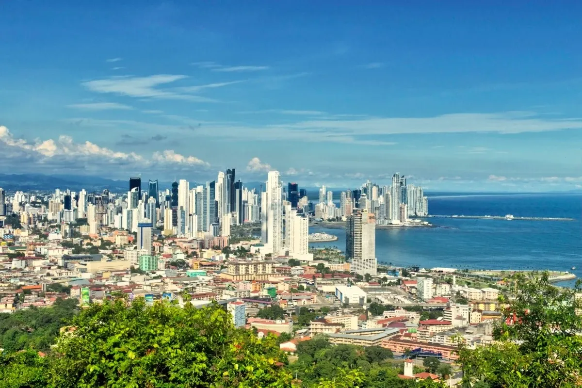 Panoramic view of Panama City skyline with modern skyscrapers, coastal waterfront, blue ocean, and lush green hills in the foreground under a clear sunny sky.