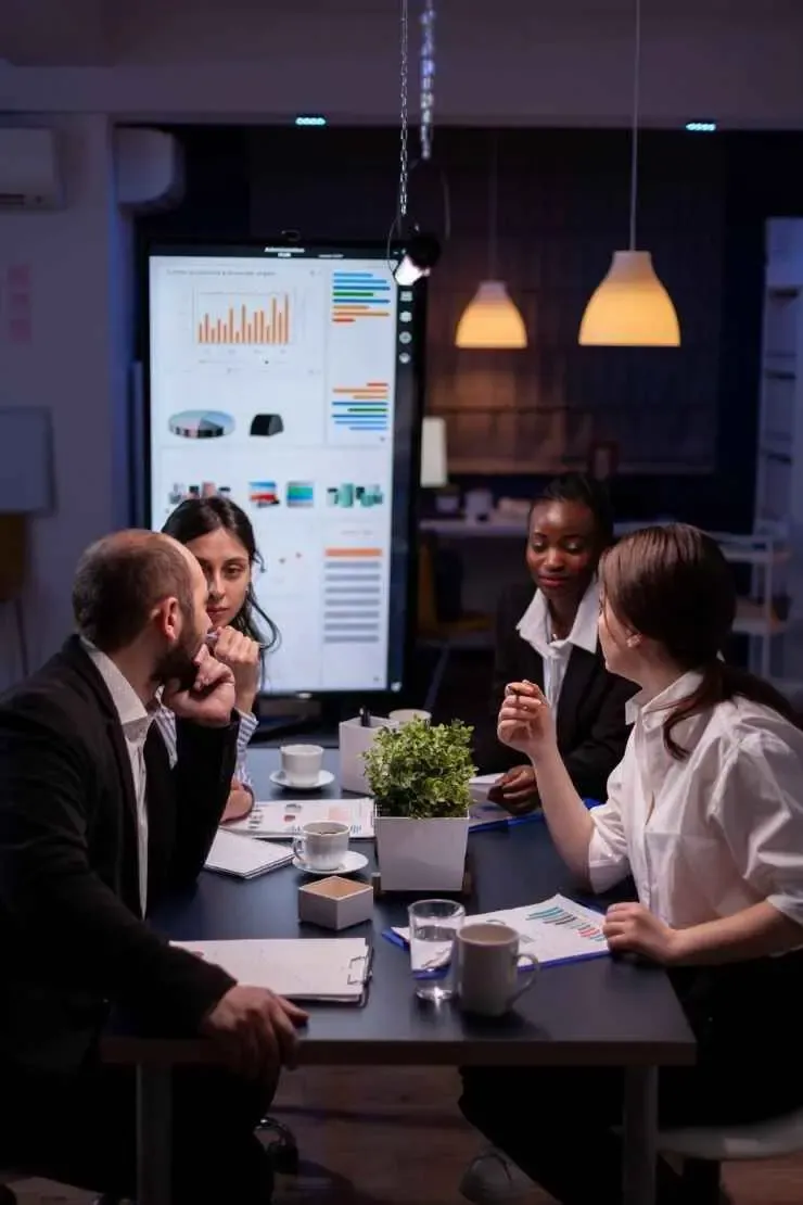 Professionals in a meeting room discussing analytics displayed on a large screen, representing business services and consulting.