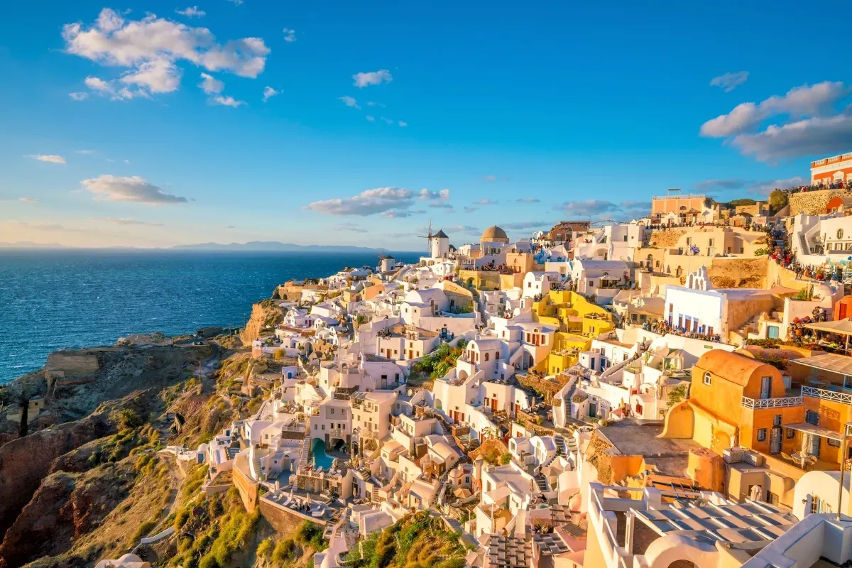 Scenic view of whitewashed hillside buildings overlooking the blue Aegean Sea in Santorini, Greece, with a bright sky and coastal cliffs.