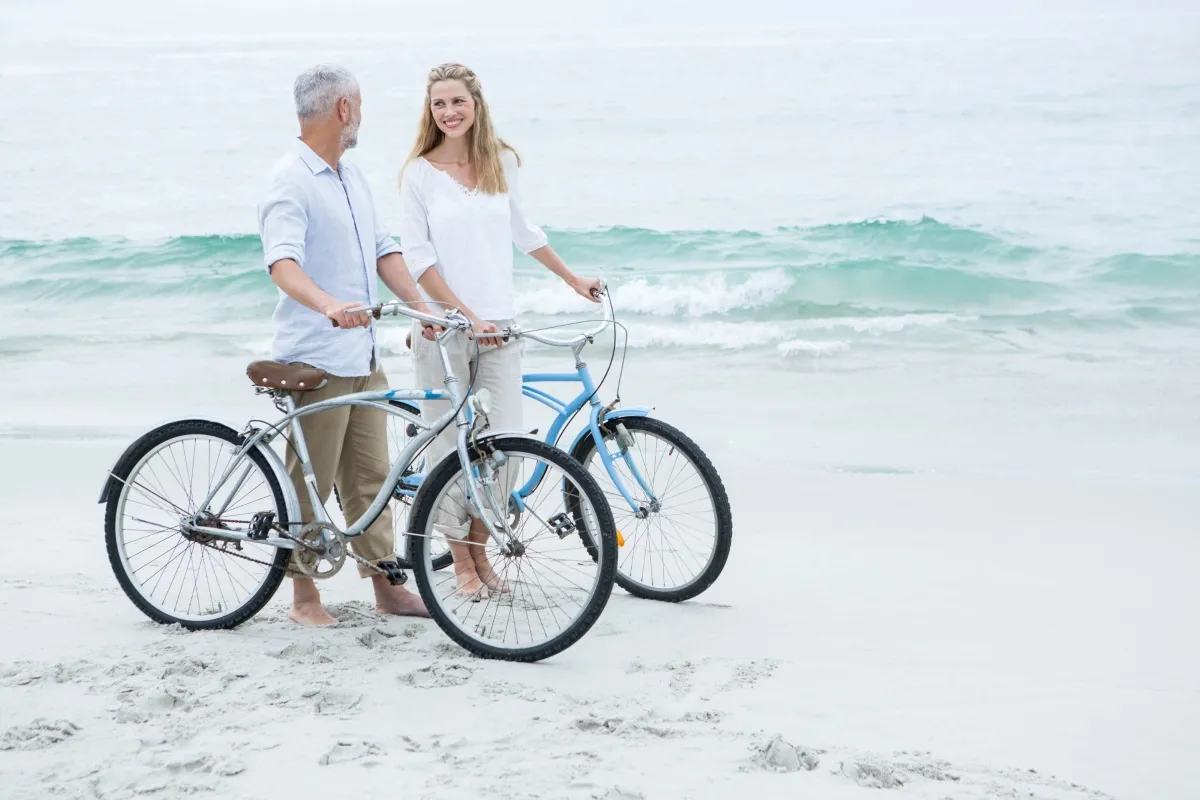 Couple standing barefoot on a sandy beach with bicycles, smiling beside ocean waves, representing retirement lifestyle, travel freedom, wellness, and leisure by the sea.