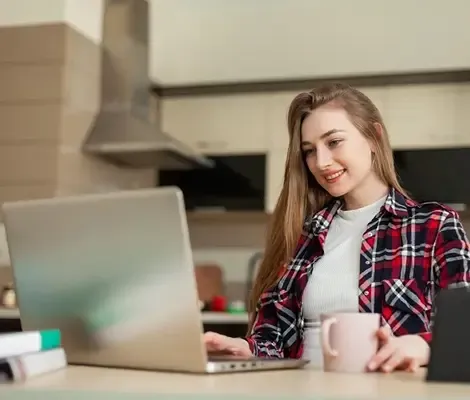 Young woman using a laptop at her kitchen table while holding a mug, appearing focused and engaged.