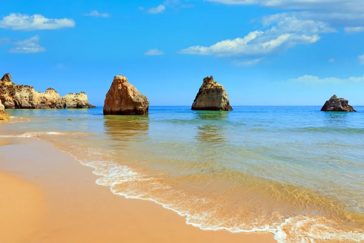 Sunny beach with clear turquoise water, golden sand, gentle waves, and rocky sea stacks under a blue sky.