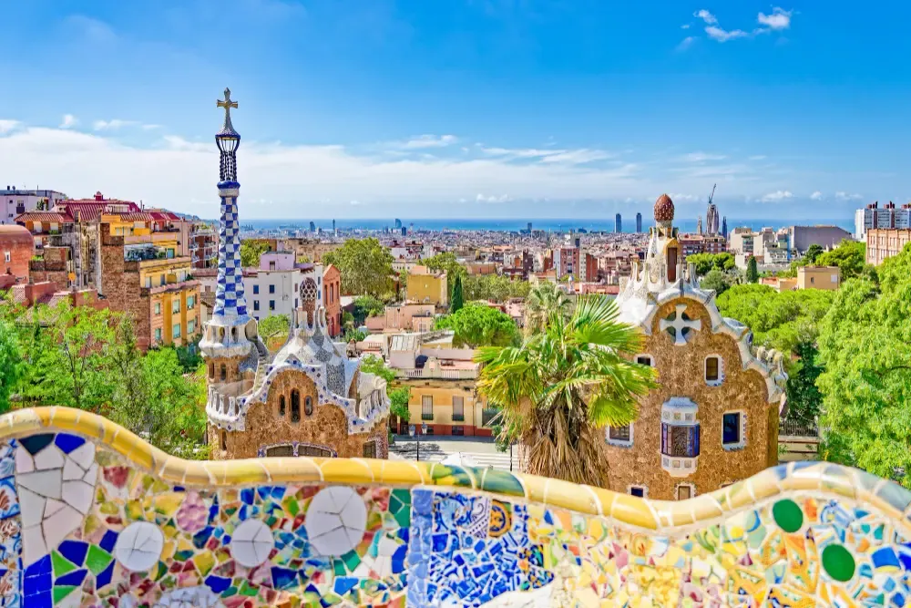 A scenic view from Park Güell featuring Antoni Gaudí’s colorful mosaic-tiled pavilions overlooking the Barcelona city skyline and the Mediterranean Se