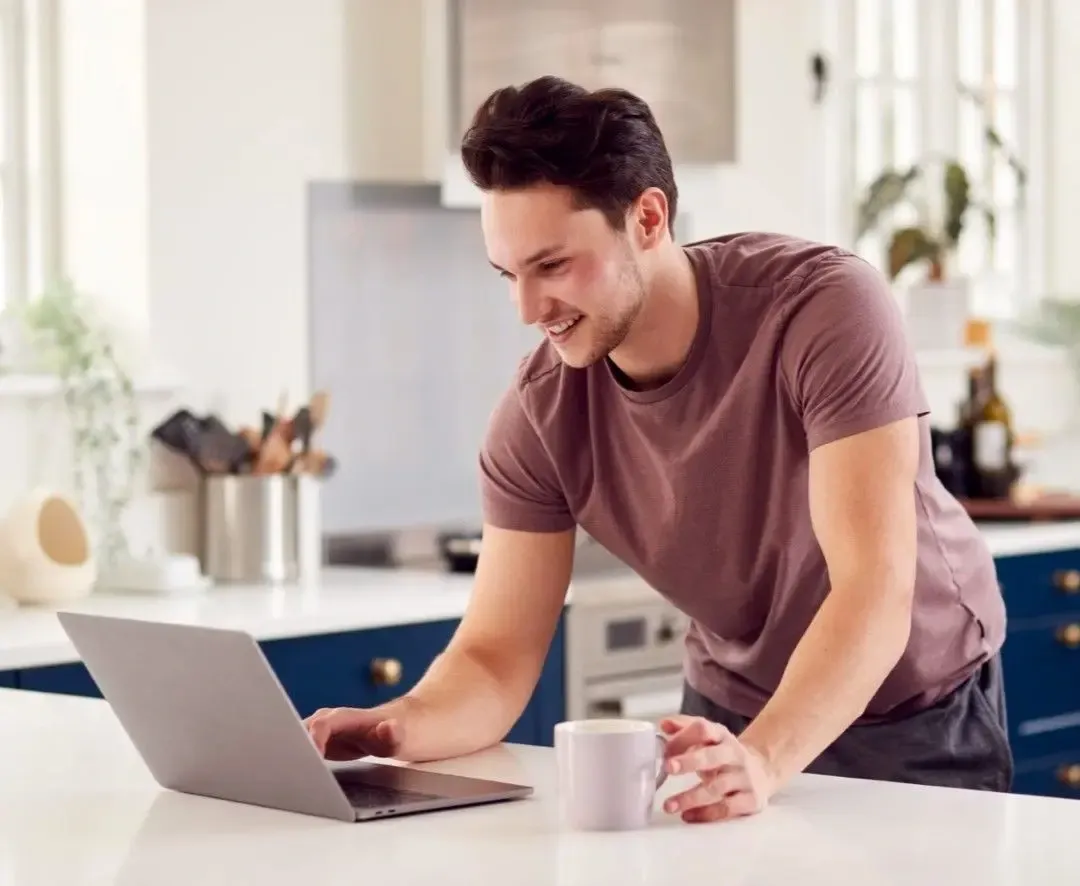 Smiling man using a laptop in a modern kitchen while holding a coffee mug