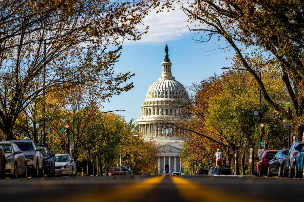 US Capitol building viewed from a wide Washington, D.C. street lined with cars, traffic lights, buildings, and leafless trees under a blue sky.