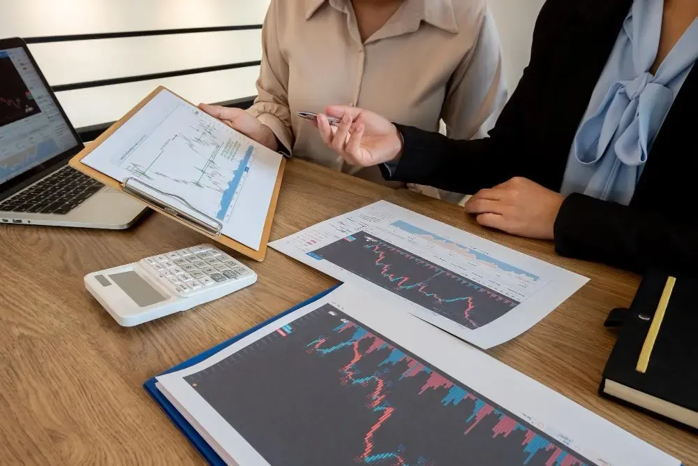 Colleagues discussing market trends using printed charts, a calculator, and a laptop in a meeting setting.