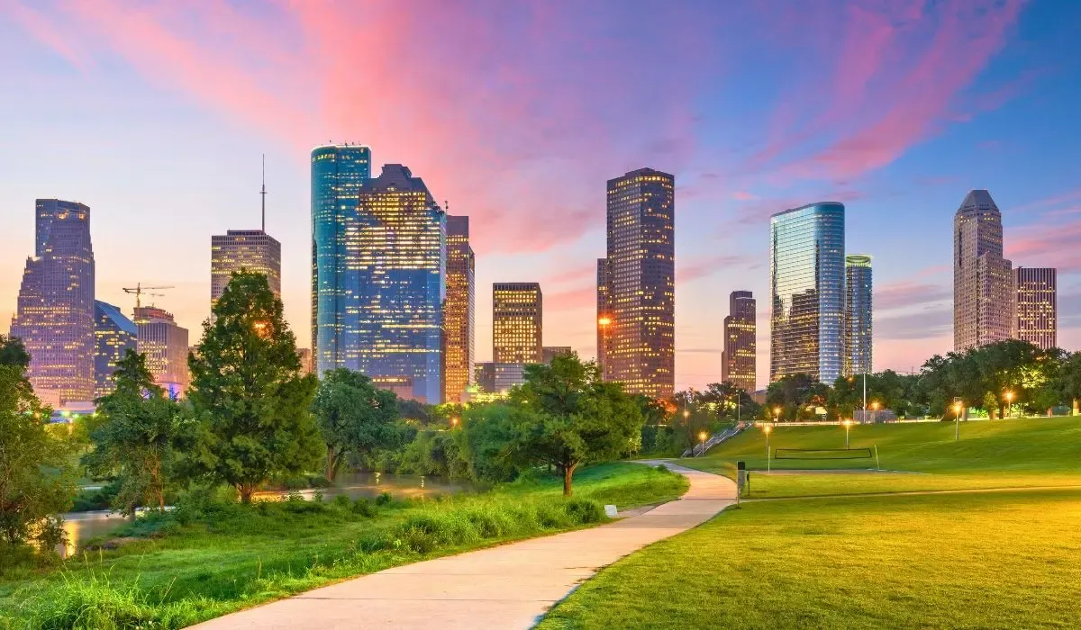 A cityscape of Houston, Texas, featuring a mix of modern skyscrapers and green spaces under a bright, sunny sky.​