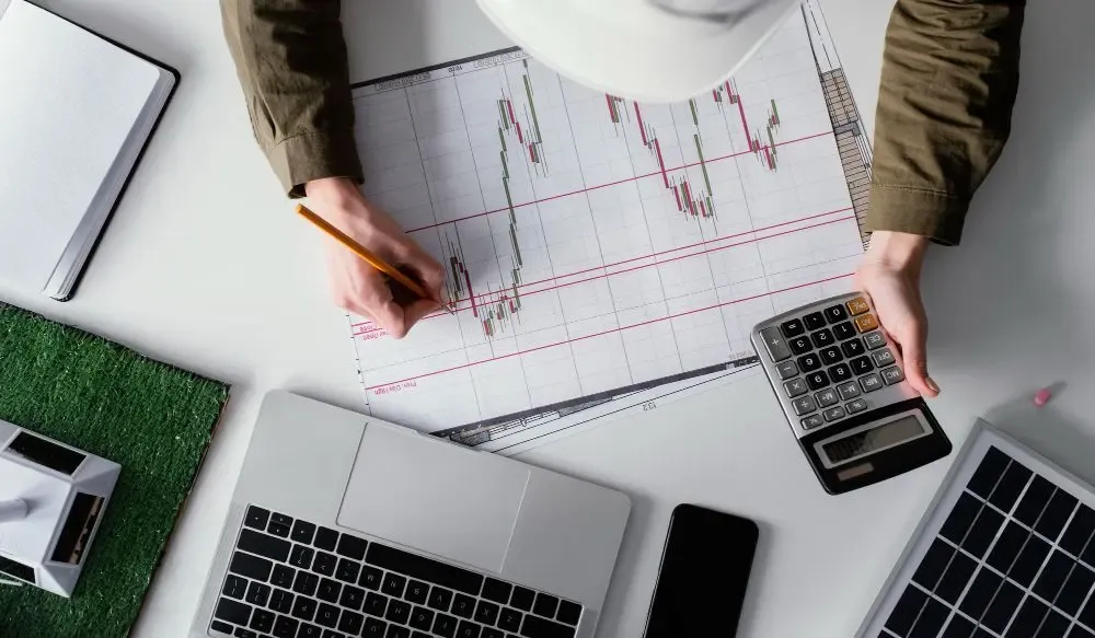 Financial analyst drawing trend lines on a printed chart with a pencil, surrounded by a laptop, calculator, and notepad.