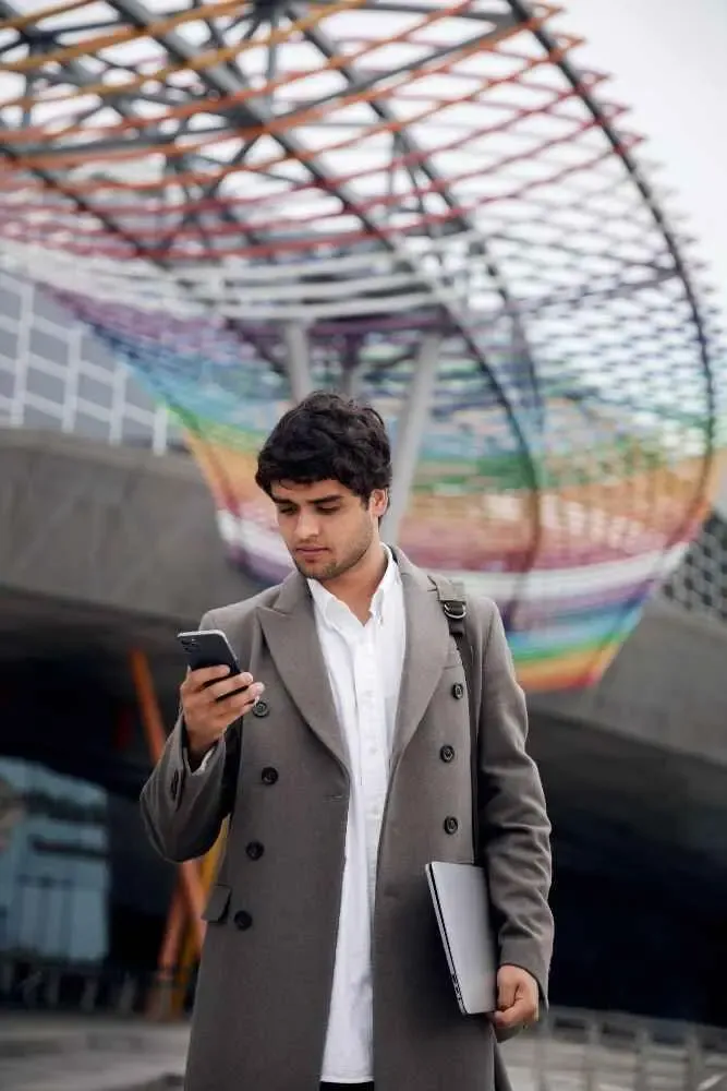 Young professional checking a mobile phone while holding a laptop in a modern urban setting, representing freelance and gig work.