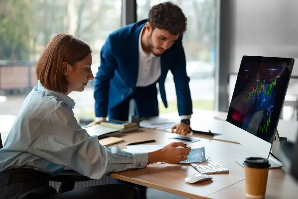 Business professionals collaborating over real-time currency charts on a desktop monitor in a modern office setting.