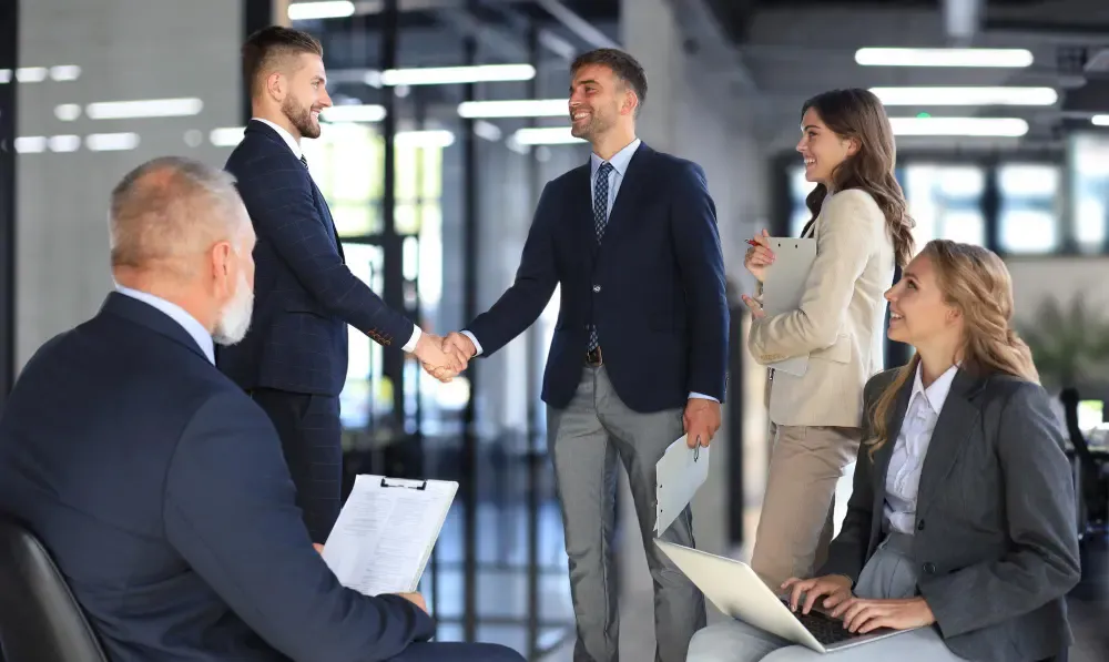 Two professionals shaking hands in a collaborative office setting with smiling colleagues.