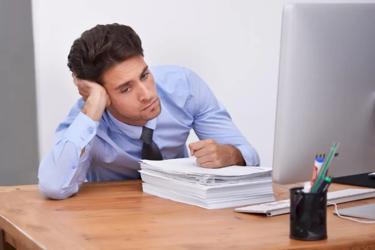 Business professional looking stressed while reviewing a large stack of paperwork at a desk, highlighting manual accounts receivable processing challenges.