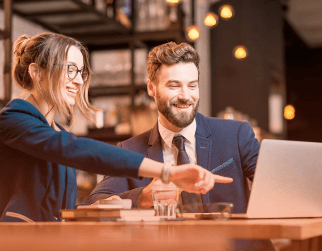 Two business professionals in formal attire collaborating and pointing at a laptop screen in a modern café setting.