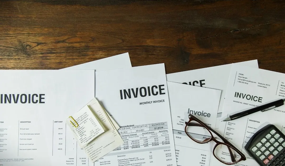 Office desk with multiple printed invoices, calculator, eyeglasses, pen, and receipts on a wooden table, representing business accounting, bill payments, bookkeeping, and expense management.