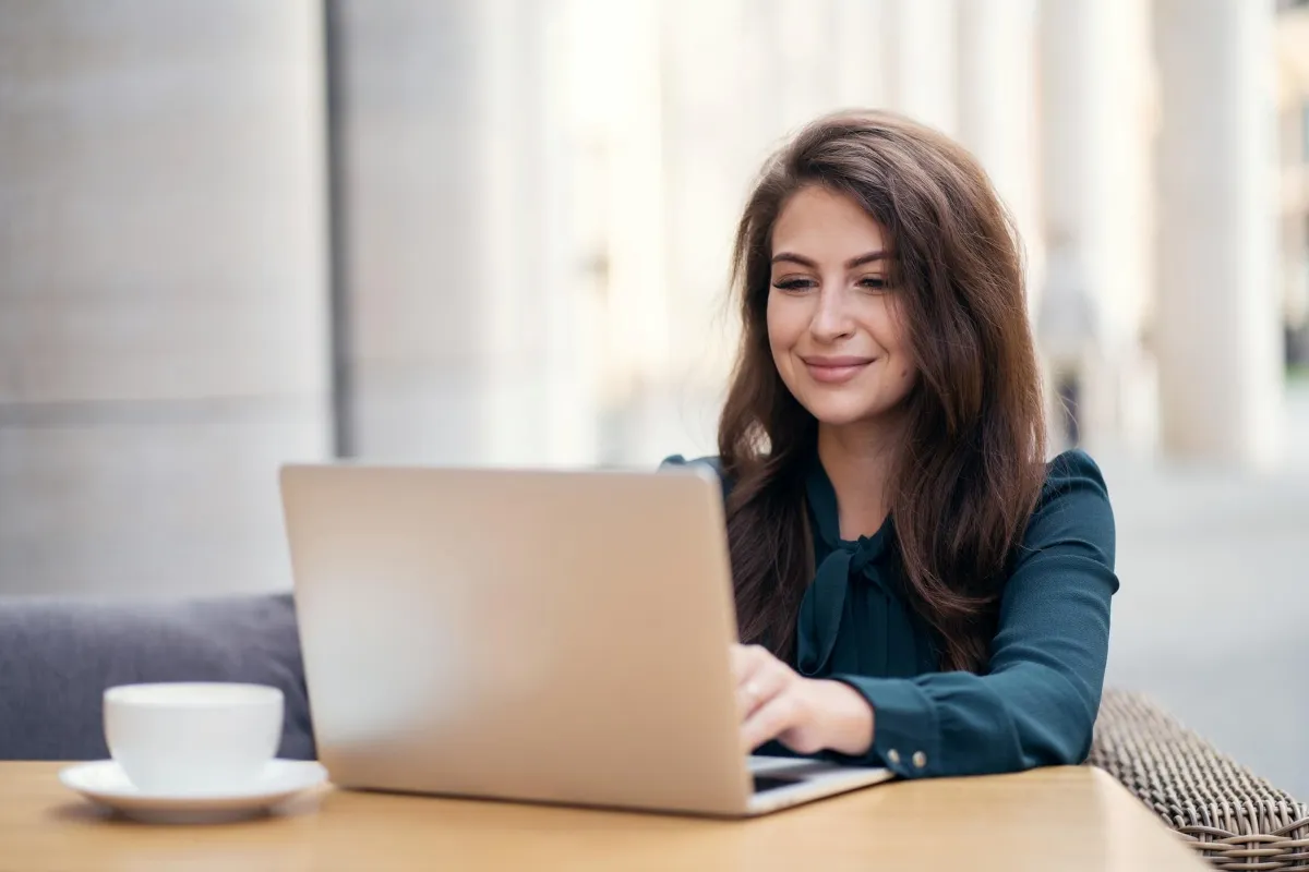 Woman working on a laptop at an outdoor café table, smiling while typing, with a coffee cup beside her and soft urban architecture in the background.