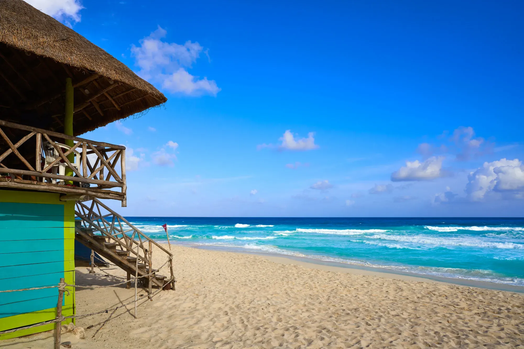 Sunny tropical beach with turquoise ocean waves, blue sky, sandy shoreline, and a colorful thatched-roof hut on the left.