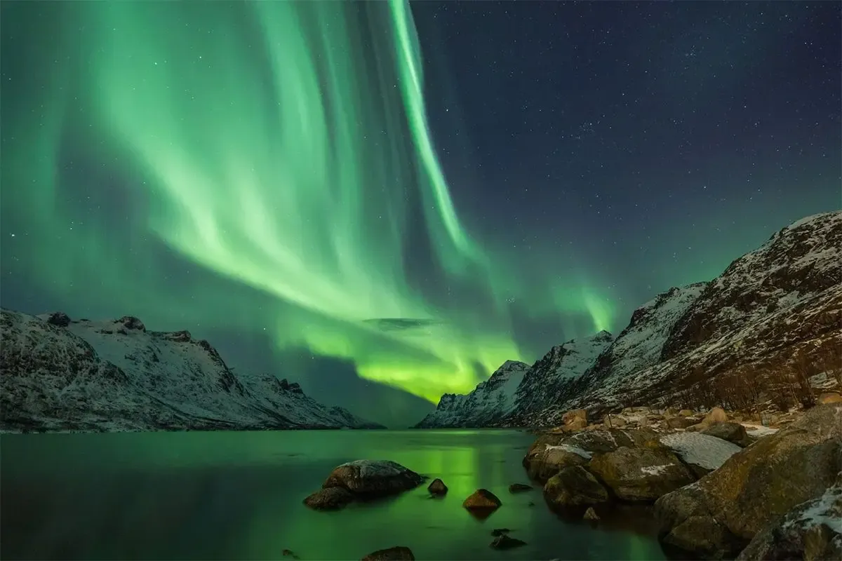 Northern lights over snow-covered Norwegian fjords with mountain peaks reflecting in the water at night.