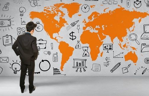 A businessman standing in front of a wall with an orange world map surrounded by hand-drawn business icons, including graphs, currency symbols, documents, and technology tools, symbolizing global business strategy and expansion.