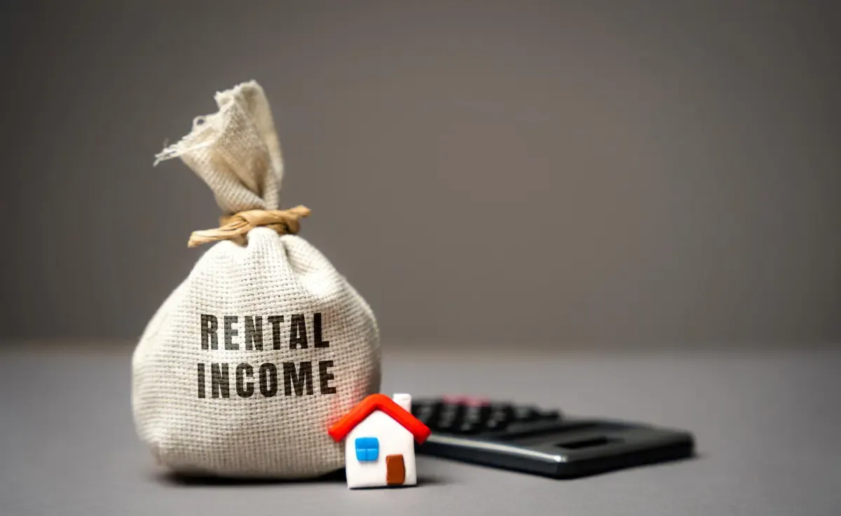 Canvas bag labeled rental income beside miniature house and calculator representing property rental earnings and financial planning.