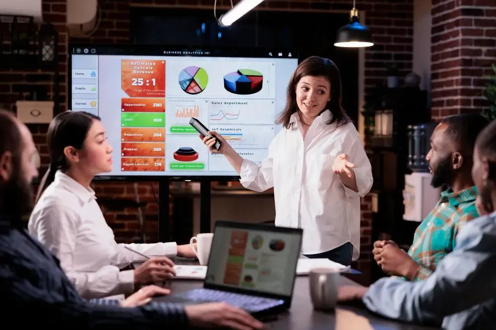 Businesswoman presenting weekly currency performance data on a large screen to colleagues during a strategy meeting.