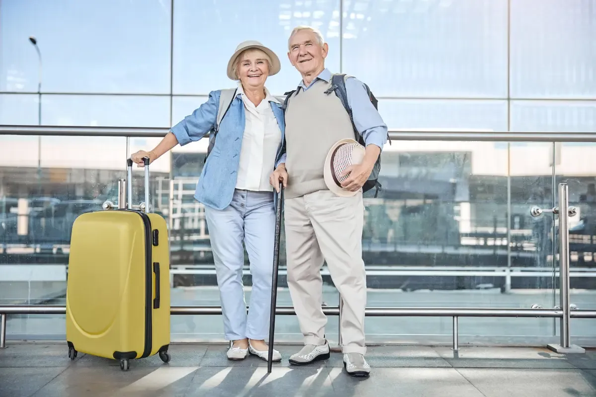 Smiling senior couple standing at an airport terminal with travel backpacks and a bright yellow suitcase, ready for an overseas retirement trip.