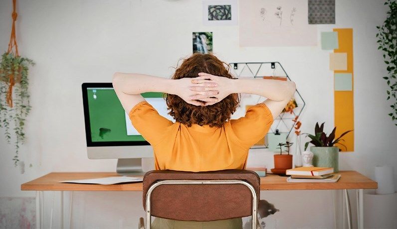 Person relaxing at home office desk, facing computer screen, surrounded by indoor plants and colorful notes on the wall.