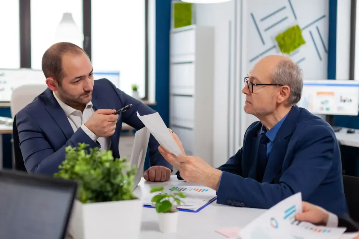 Two business professionals reviewing financial documents in an office setting, discussing reports and data related to payments or financial planning.