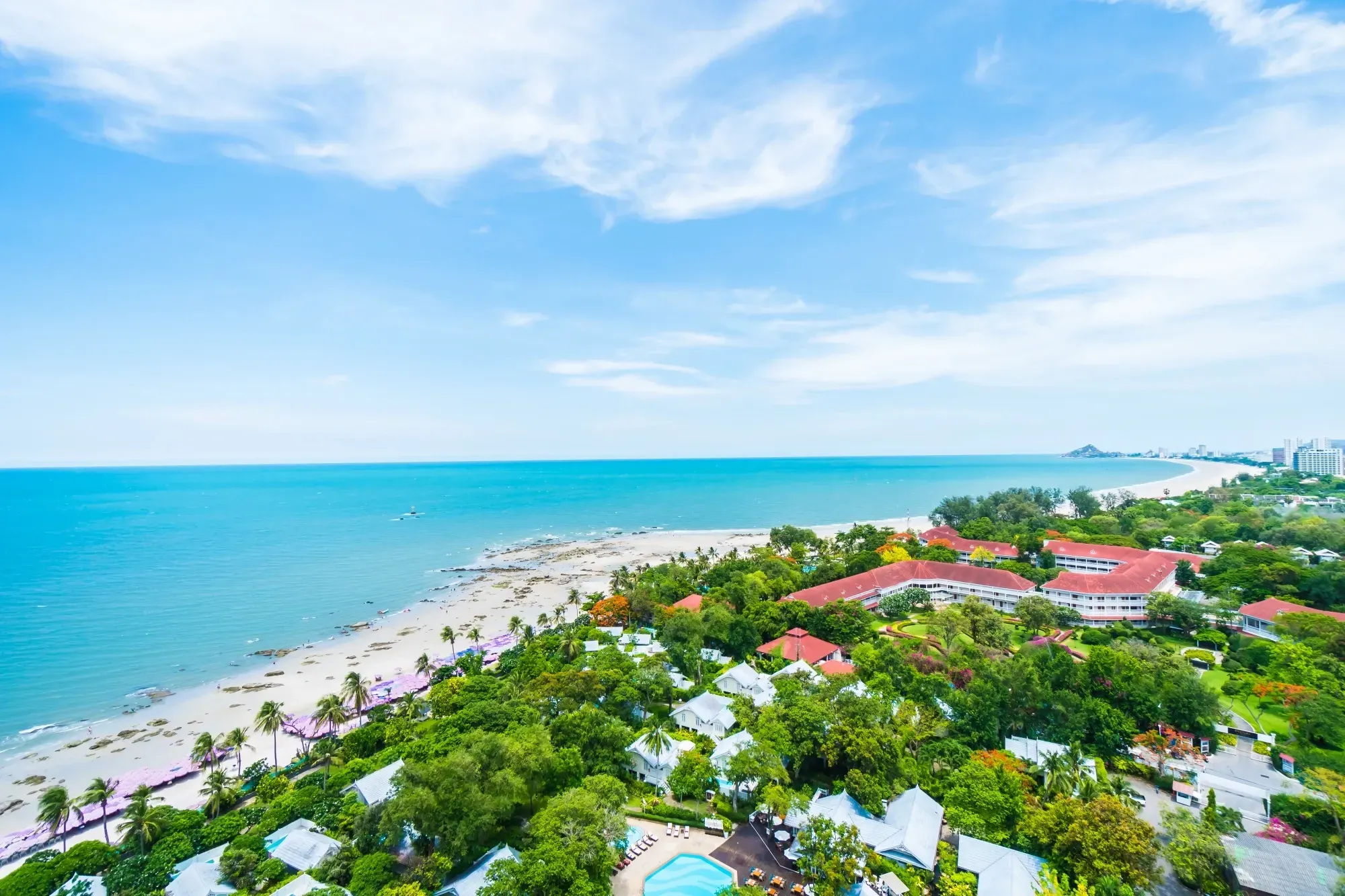 Aerial view of a tropical beach with turquoise ocean, white sand, and lush green coastal resort buildings under a blue sky.