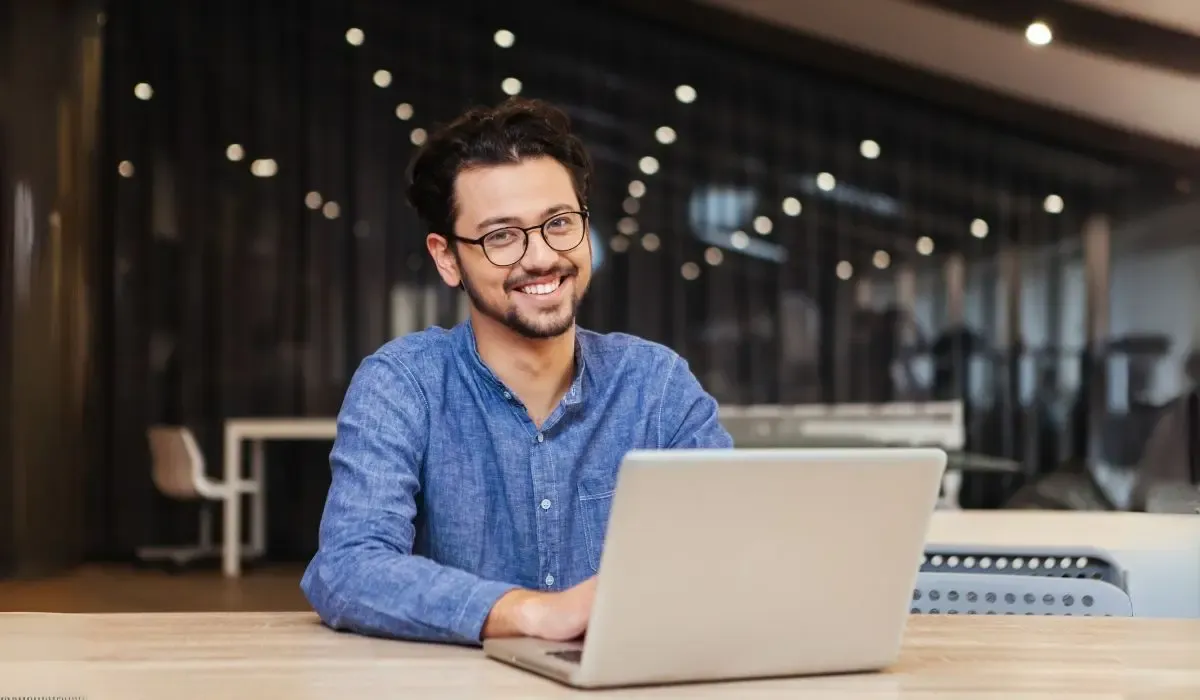 An image of a young professional man wearing glasses and a blue shirt, smiling confidently while working on a laptop in a modern office environment.