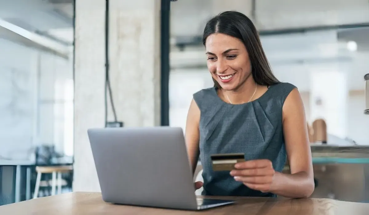 Smiling woman holding a credit card while using a laptop, representing online payments or money transfers in a modern office setting.