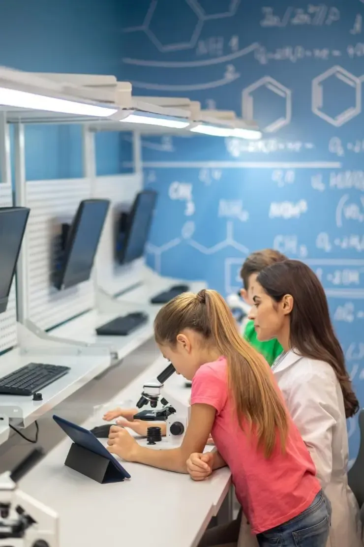 Students using digital tablets in a modern classroom equipped with computers and microscopes, representing educational technology.