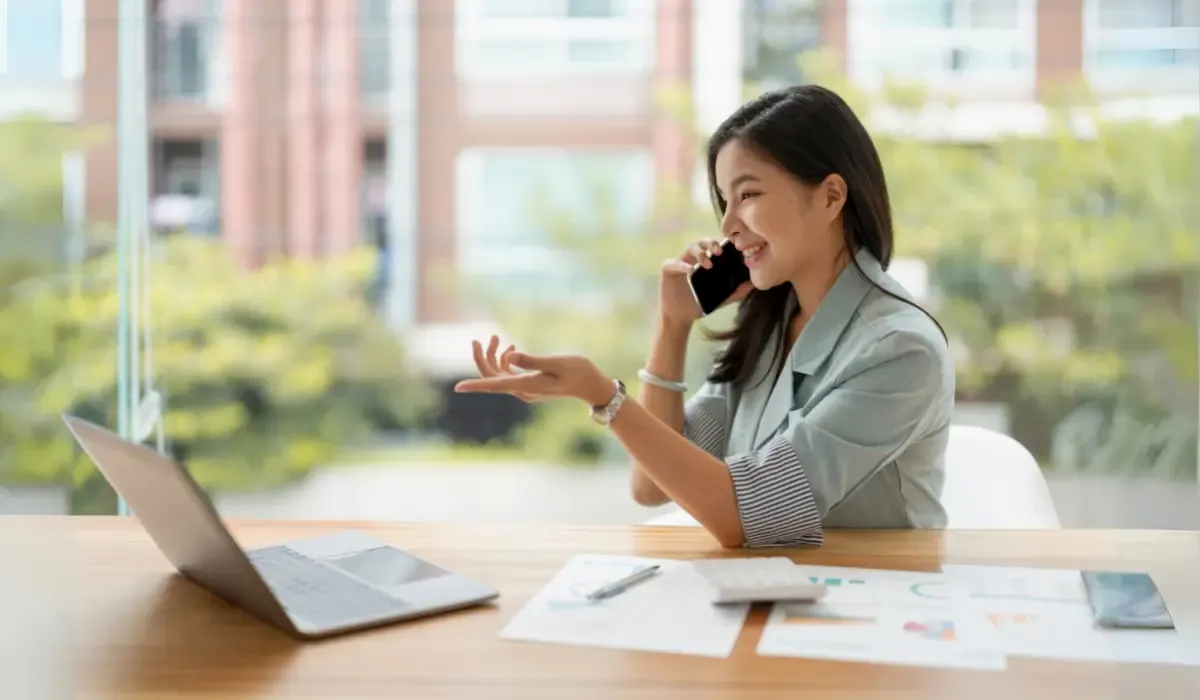 A businesswoman sitting at her desk, holding a phone to her ear while gesturing with her hand, with a laptop and documents on the table in a bright office setting.
