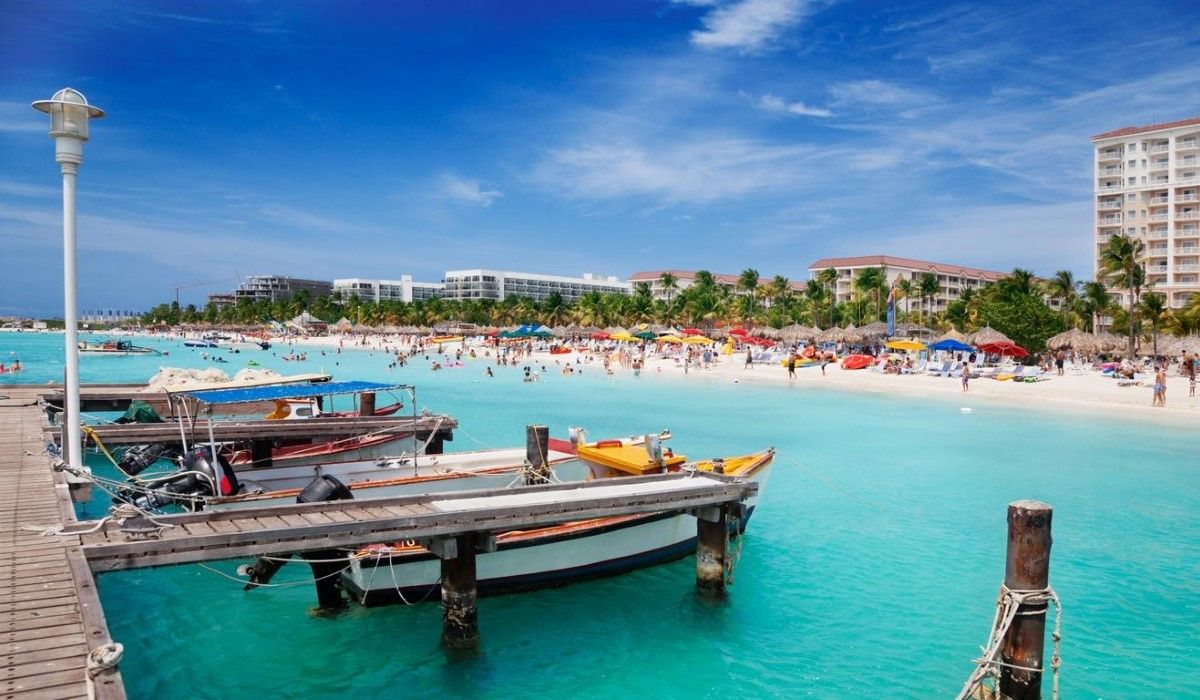 A scenic view of boats docked at a wooden pier along a turquoise beach in Aruba, with a vibrant crowd enjoying the white sandy shore.