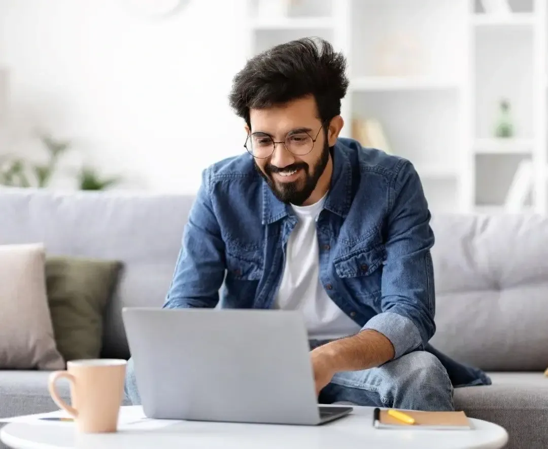 Man sitting on a couch and smiling while working on a laptop at home with a coffee mug nearby