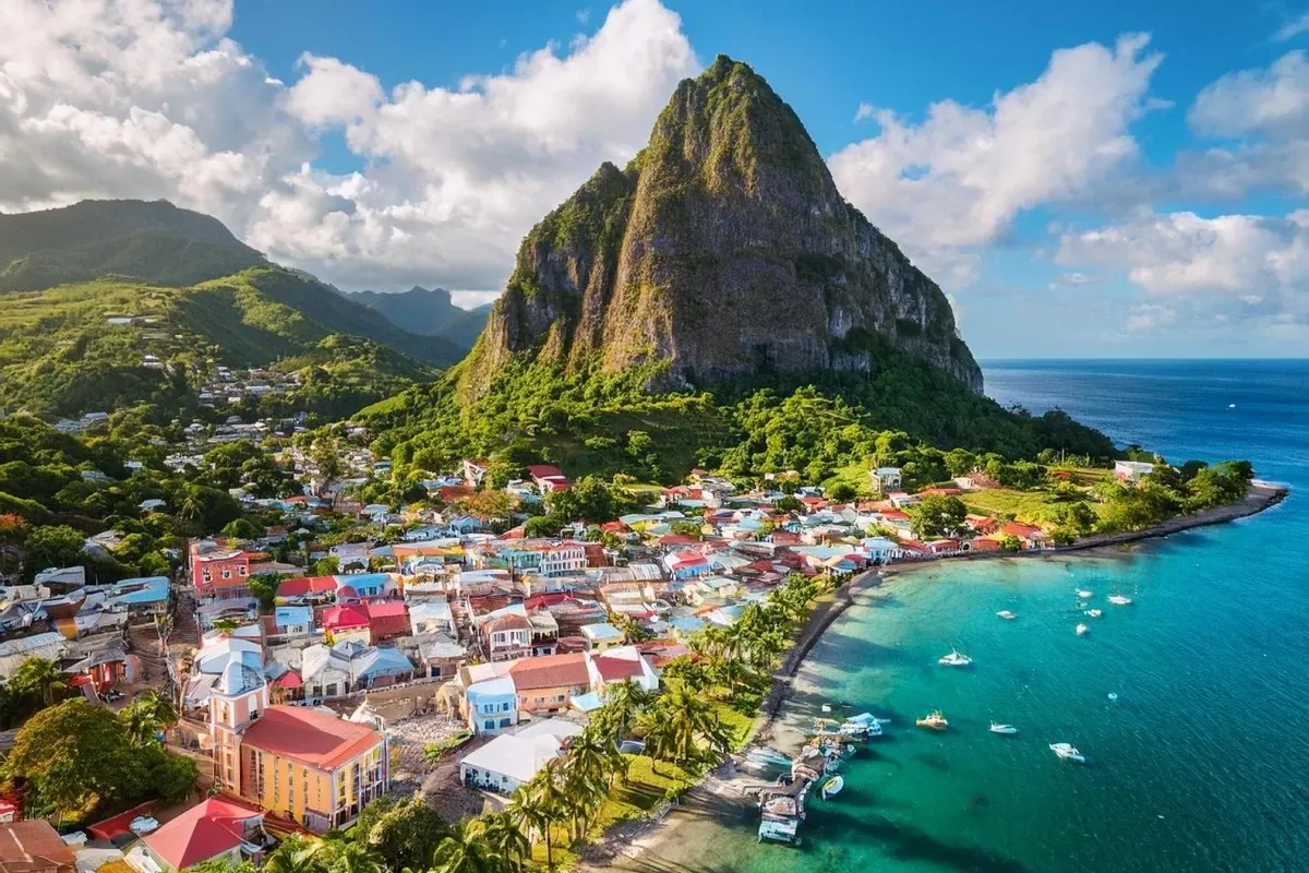 Aerial view of a colourful coastal town beside turquoise waters and dramatic green mountains, showcasing tropical island scenery, seaside living, travel, and natural beauty.