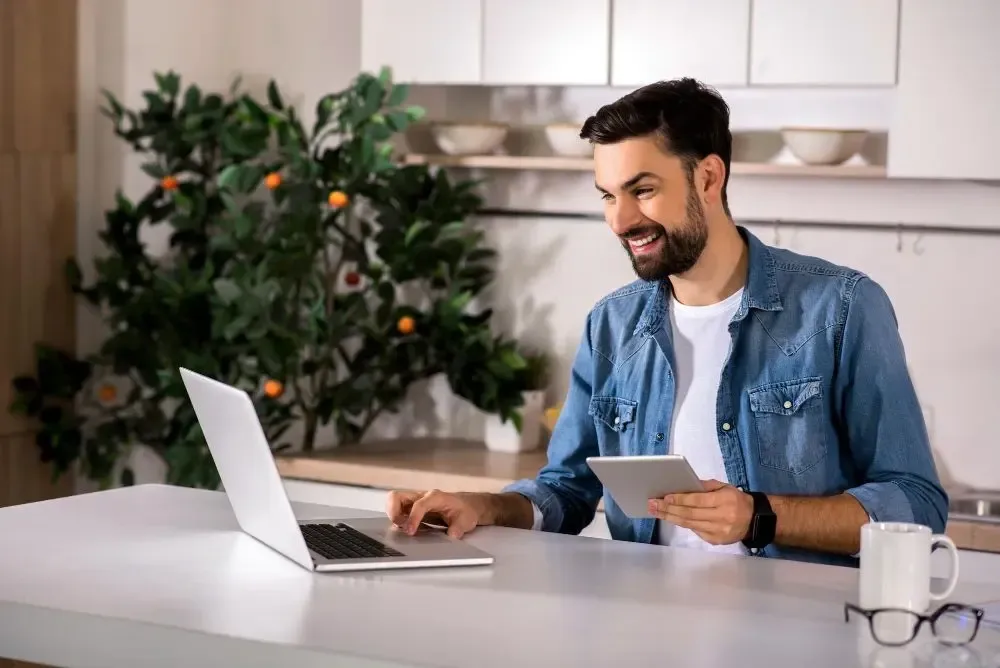 Smiling man working on a laptop and holding a tablet in a modern kitchen