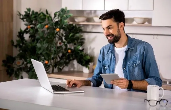 Smiling man using a laptop and tablet at home, representing convenient and secure online money transfers for international mortgage payments.
