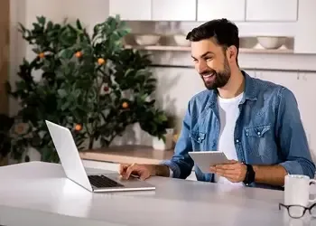 Smiling man using a laptop and tablet at home, representing convenient and secure online money transfers for international mortgage payments.