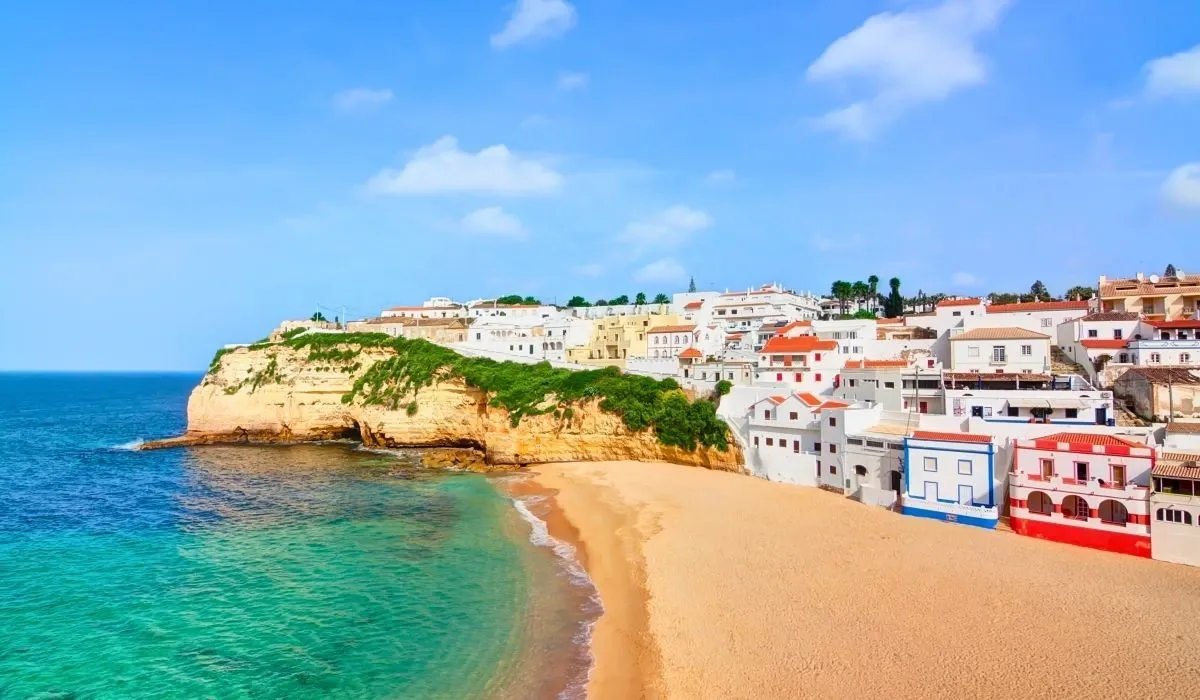 Charming whitewashed buildings perched on cliffs above a golden beach in the Algarve region of Portugal, with clear turquoise waters.