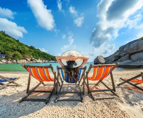 Person relaxing on a beach chair facing the ocean, surrounded by colorful loungers on a tropical beach