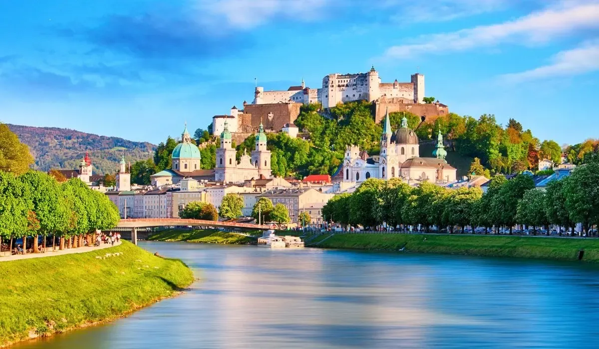 A panoramic view of Vienna, Austria, showcasing historic baroque architecture, including the Hofburg Palace, with the Alps visible in the background under a clear blue sky.