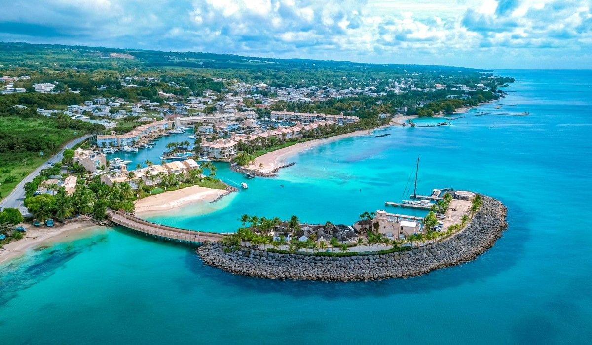 Aerial view of a luxury coastal community in Barbados featuring turquoise waters, white sand beaches, upscale villas, and a marina with docked sailboats.