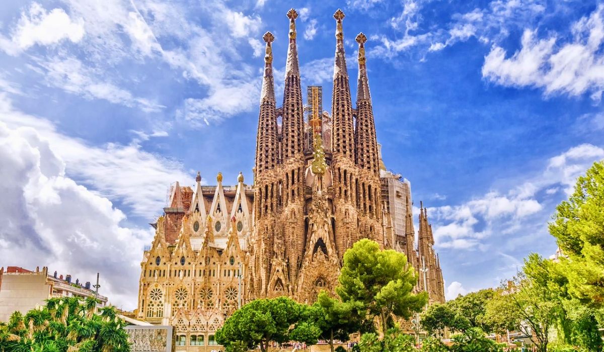 A landscape view of Basílica i Temple Expiatori de la Sagrada Família in Barcelona with trees in the foreground and blue skies in the backdrop.