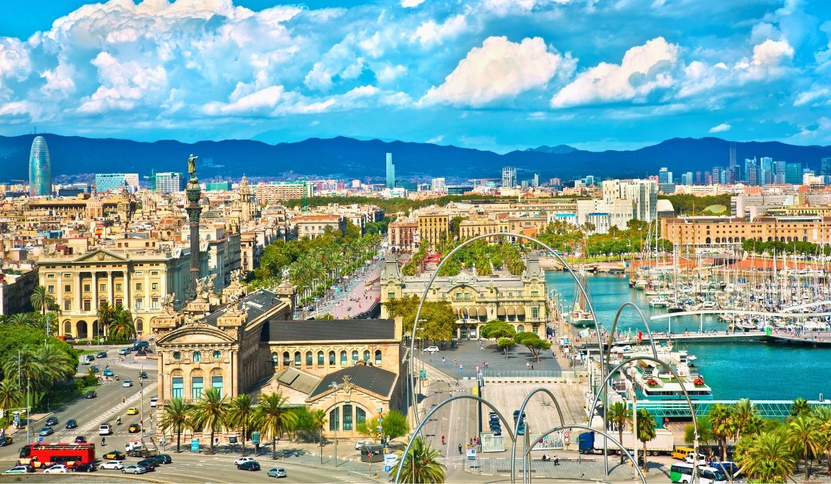 A panoramic view of Barcelona’s waterfront, showcasing the Columbus Monument, marina filled with yachts, historic buildings, and the city skyline backed by distant mountains.