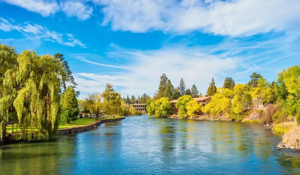 A picturesque image of Bend, Oregon, featuring the Deschutes River, pine forests, and snow-capped mountains in the distance.​