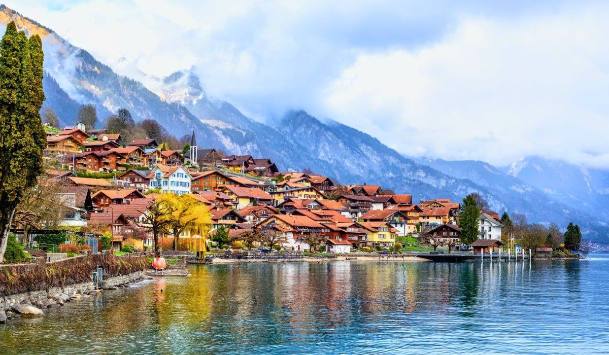 Lakeside village in Switzerland with chalet-style homes and snow-capped alpine mountains in the background.