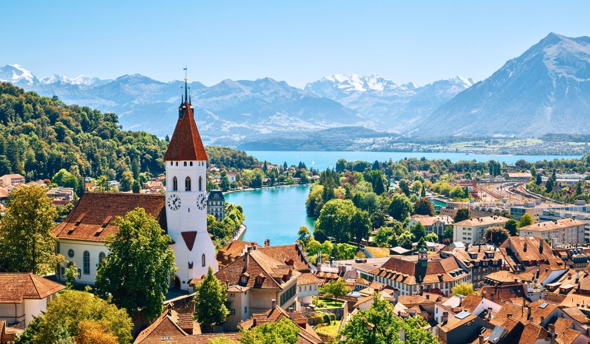 Panoramic view of Bern, Switzerland, with a clock tower and rooftops overlooking Lake Thun and the Swiss Alps.