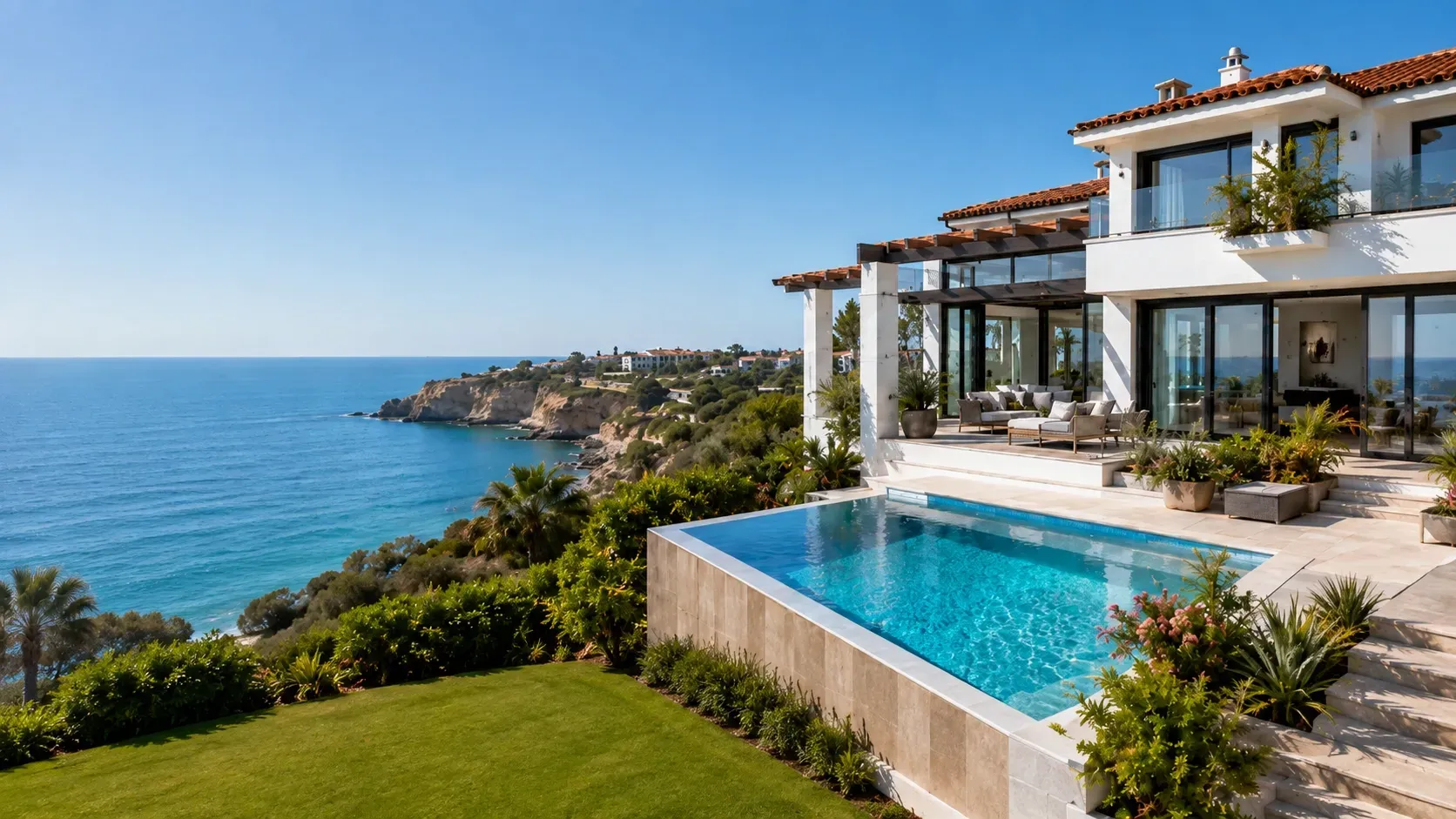 Modern white coastal vacation villa with an infinity pool overlooking the blue sea and rocky shoreline under a clear sky.