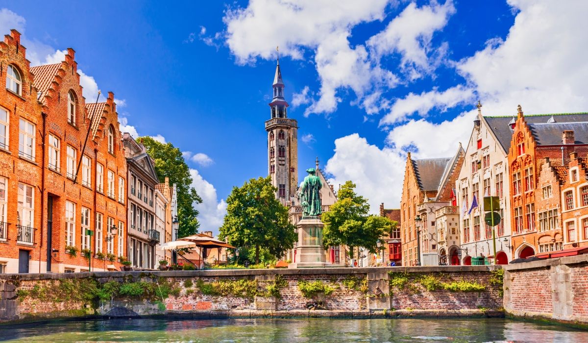 Charming canal-side view of Bruges, Belgium, with colorful medieval buildings and a tall clock tower under a bright blue sky.