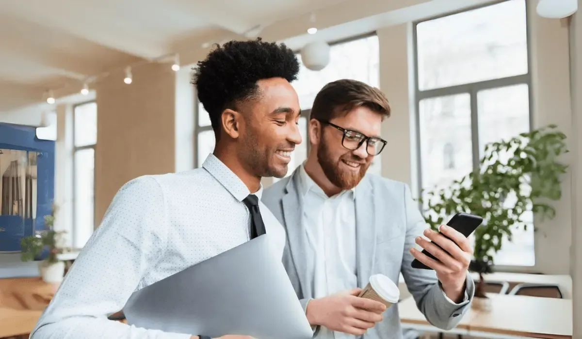 Two smiling businessmen in a modern office reviewing something on a smartphone while holding coffee and a laptop.
